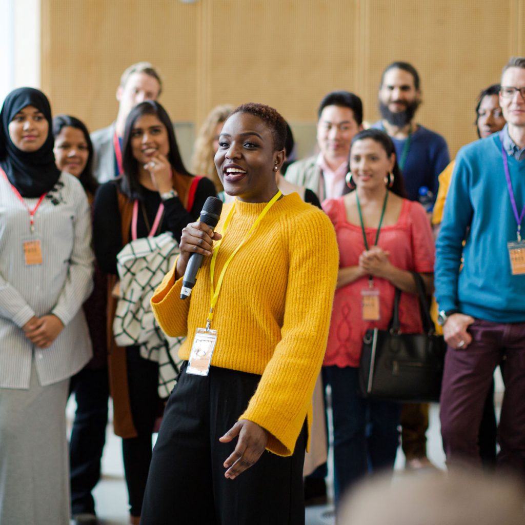An image of a speaker in the middle of a crowd, addressing attendees at an event